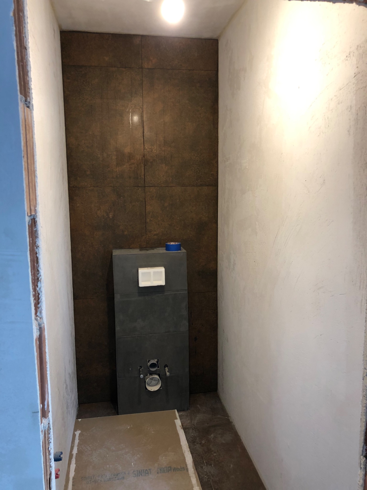 Wnętrze łazienki w trakcie prac: ściany częściowo wykończone gładzią, ciemnobrązowe płytki na ścianie z zamontowanym stelażem podtynkowym WC, widoczna płyta karton-gips na podłodze.