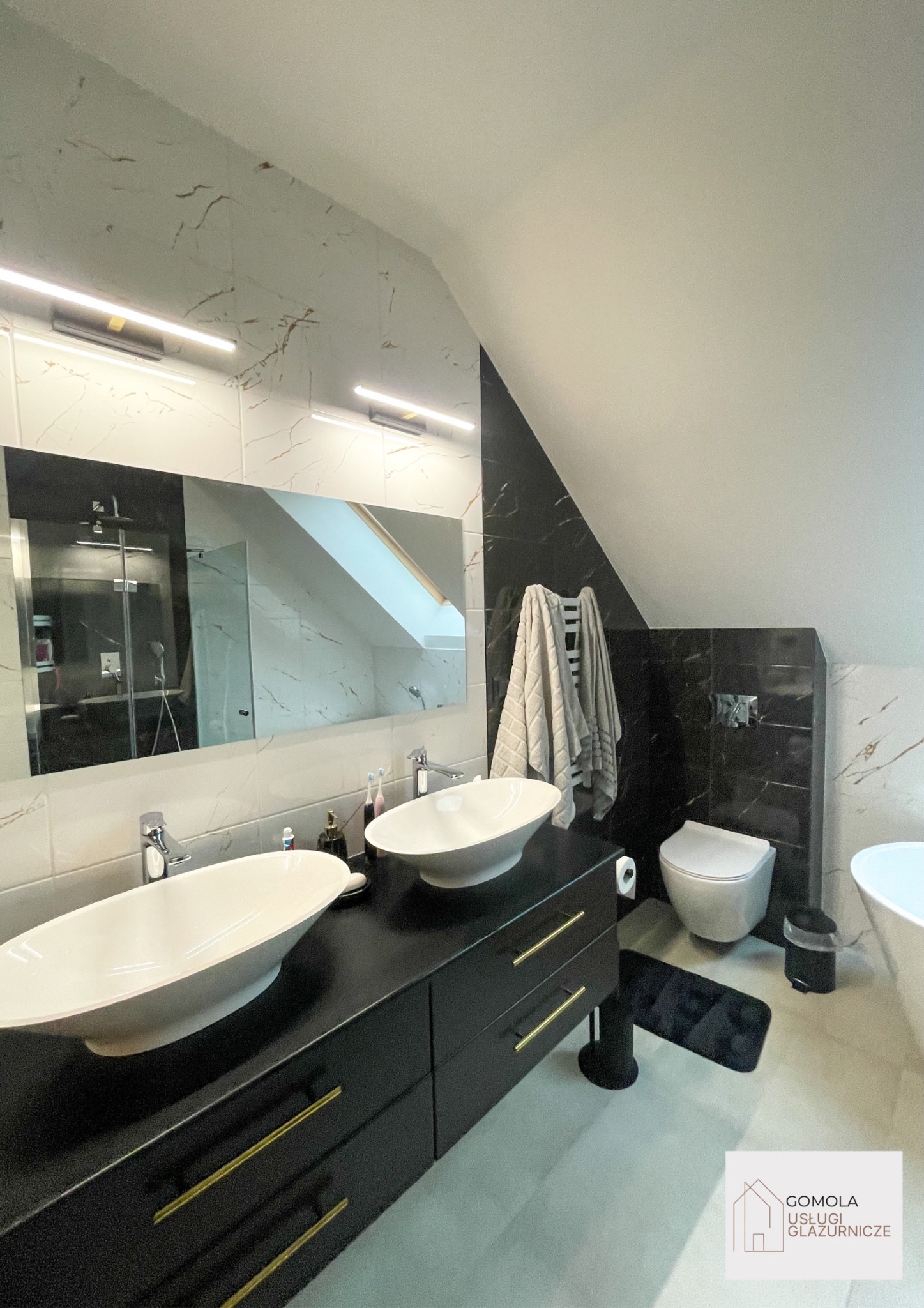 Nowoczesna łazienka na poddaszu z dwoma owalnymi umywalkami nablatowymi, czarną szafką z złotymi uchwytami, marmurowymi płytkami na ścianach i toaletą w tle.
