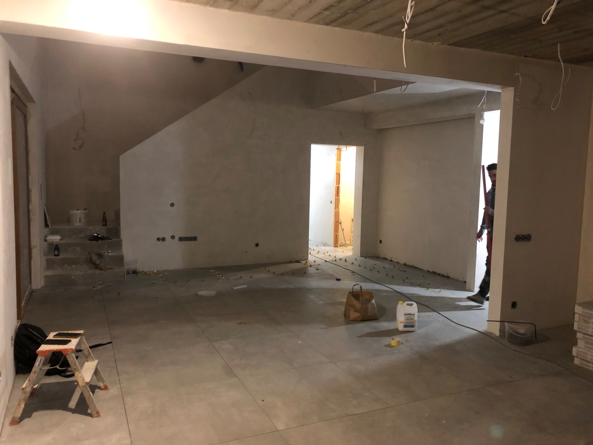 Wnętrze w trakcie prac wykończeniowych, z ułożonymi płytkami podłogowymi, schodami w tle i widocznymi narzędziami oraz materiałami budowlanymi, w tym poziomica na schodach i wiadro z zaprawą w rogu...