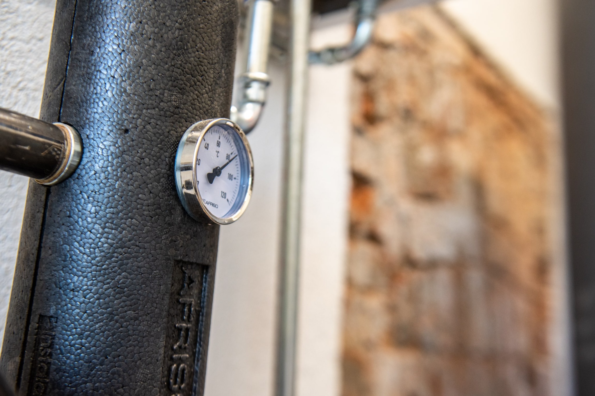 Izolowany rurociąg z zamontowanym termometrem Afriso, widoczny na tle ściany z cegieł.