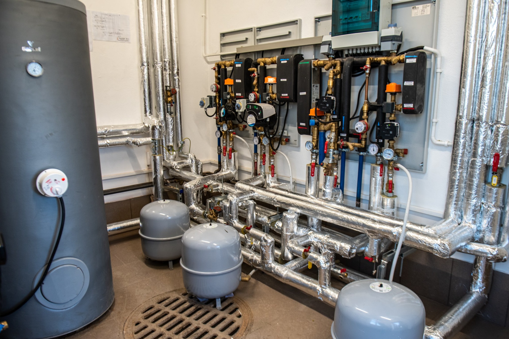 Węzeł cieplny z licznymi rurami w izolacji termicznej, zbiornikami wyrównawczymi i rozbudowaną szafą sterowniczą z manometrami, zamontowany w pomieszczeniu technicznym.