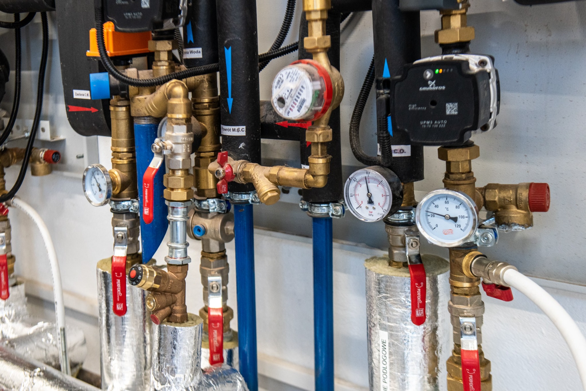 Węzeł cieplny z licznikiem ciepła, manometrami, zaworami z czerwonymi uchwytami i pompą obiegową Grundfos UPM3 Auto, zamontowany na rurach izolowanych folią aluminiową, z opisami 'Zasilanie C.M.'...