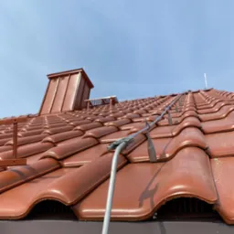 Czerwona dachówka z zainstalowaną instalacją odgromową, widok z dołu na błękitne niebo. Metalowy komin w tle. Specyficzne mocowania instalacji.