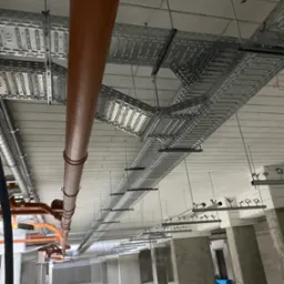 Widok z dołu na sufit z zamontowanymi korytkami kablowymi i rurami wentylacyjnymi w surowym wnętrzu budynku. Szare betonowe słupy w tle.