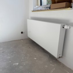 YERIS Instalacje - Biały, panelowy grzejnik zamontowany przy ścianie w surowym wnętrzu. Widoczne ślady po pracach wykończeniowych na podłodze i wystające przewody elektryczne.