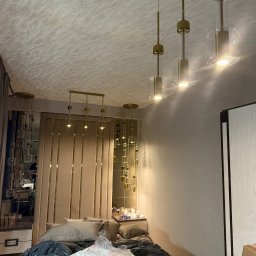 YERIS Instalacje - Nowoczesne lampy wiszące z elementami złota nad łóżkiem w sypialni. Widoczne odbicie w lustrze i elementy dekoracyjne. Oświetlenie w stylu glamour.