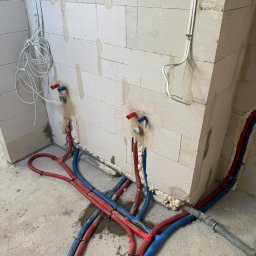YERIS Instalacje - Instalacja wodna i elektryczna w surowym wnętrzu: czerwone i niebieskie rury wodne oraz białe kable elektryczne prowadzone wzdłuż ściany z betonu komórkowego.