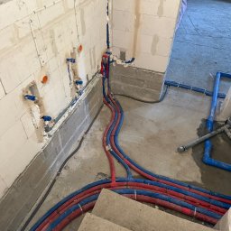 YERIS Instalacje - Wnętrze w trakcie prac hydraulicznych: czerwono-niebieskie rury prowadzone wzdłuż ściany z betonu komórkowego, z widocznymi punktami przyłączeniowymi.