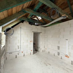 YERIS Instalacje - Surowy stan poddasza z widoczną instalacją elektryczną i wentylacyjną na ścianach z betonu komórkowego oraz drewnianą konstrukcją dachu. Widoczne kable i rury.