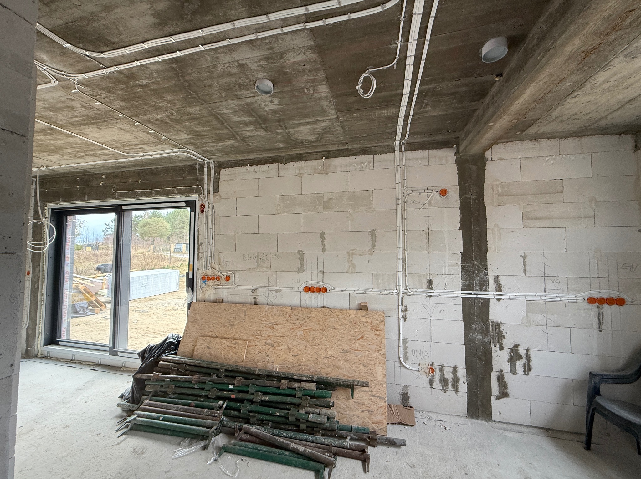 Surowy stan: białe ściany z betonu komórkowego, instalacje elektryczne w białych rurkach na suficie i ścianach. Widoczne puszki instalacyjne, okno i stemple budowlane.