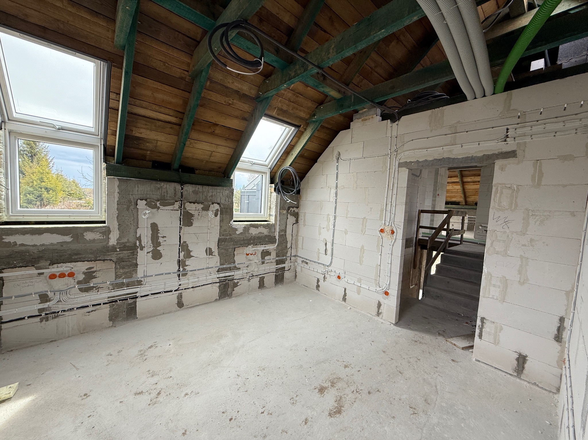 Surowy stan poddasza z instalacją elektryczną: widoczne przewody w ścianach z betonu komórkowego, okna dachowe i drewniana konstrukcja dachu.