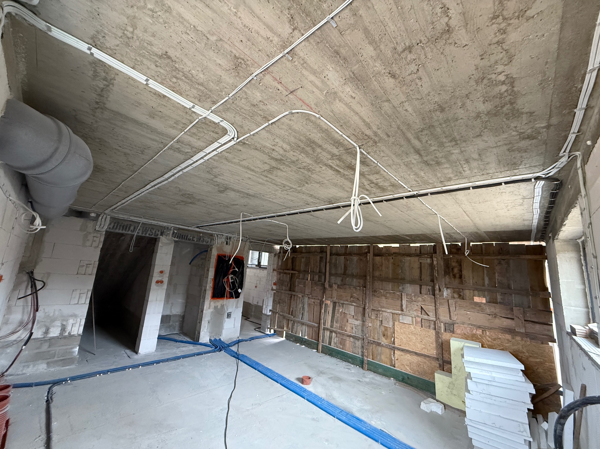 Surowy strop z instalacją elektryczną i rurami hydraulicznymi w nowym budynku. Widoczne przewody, rury w kolorze niebieskim i szare elementy wentylacyjne.