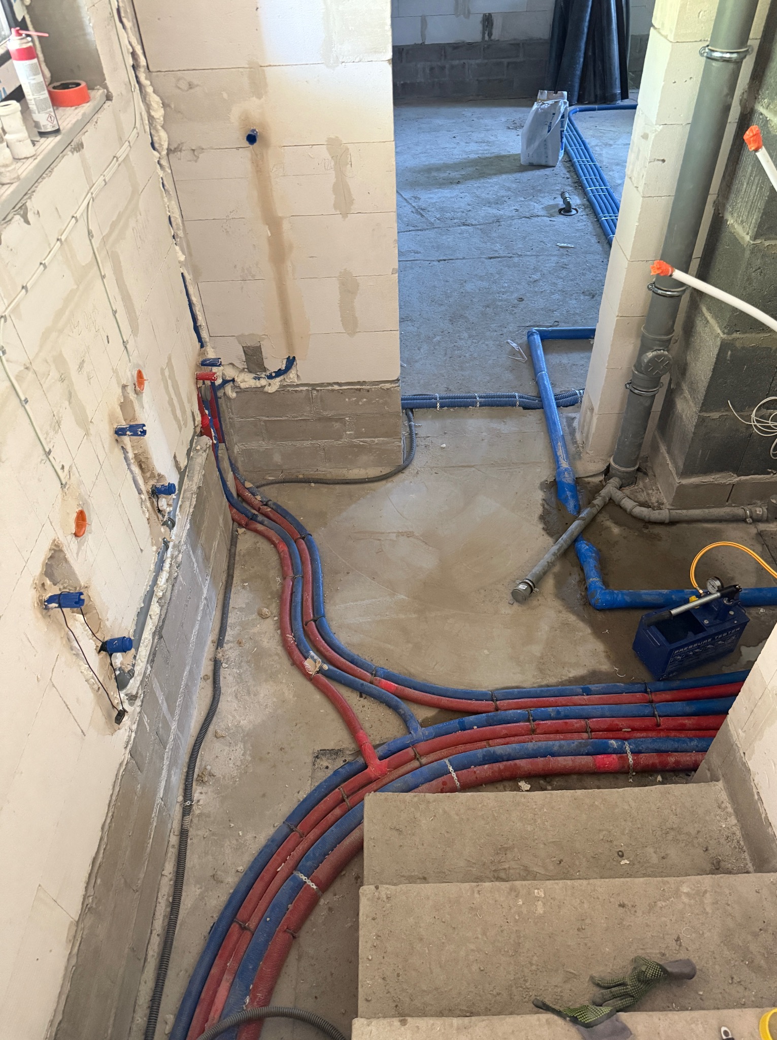 Instalacja hydrauliczna w trakcie montażu: rury czerwono-niebieskie prowadzone wzdłuż ściany, widoczne przyłącza i manometr do testowania ciśnienia.
