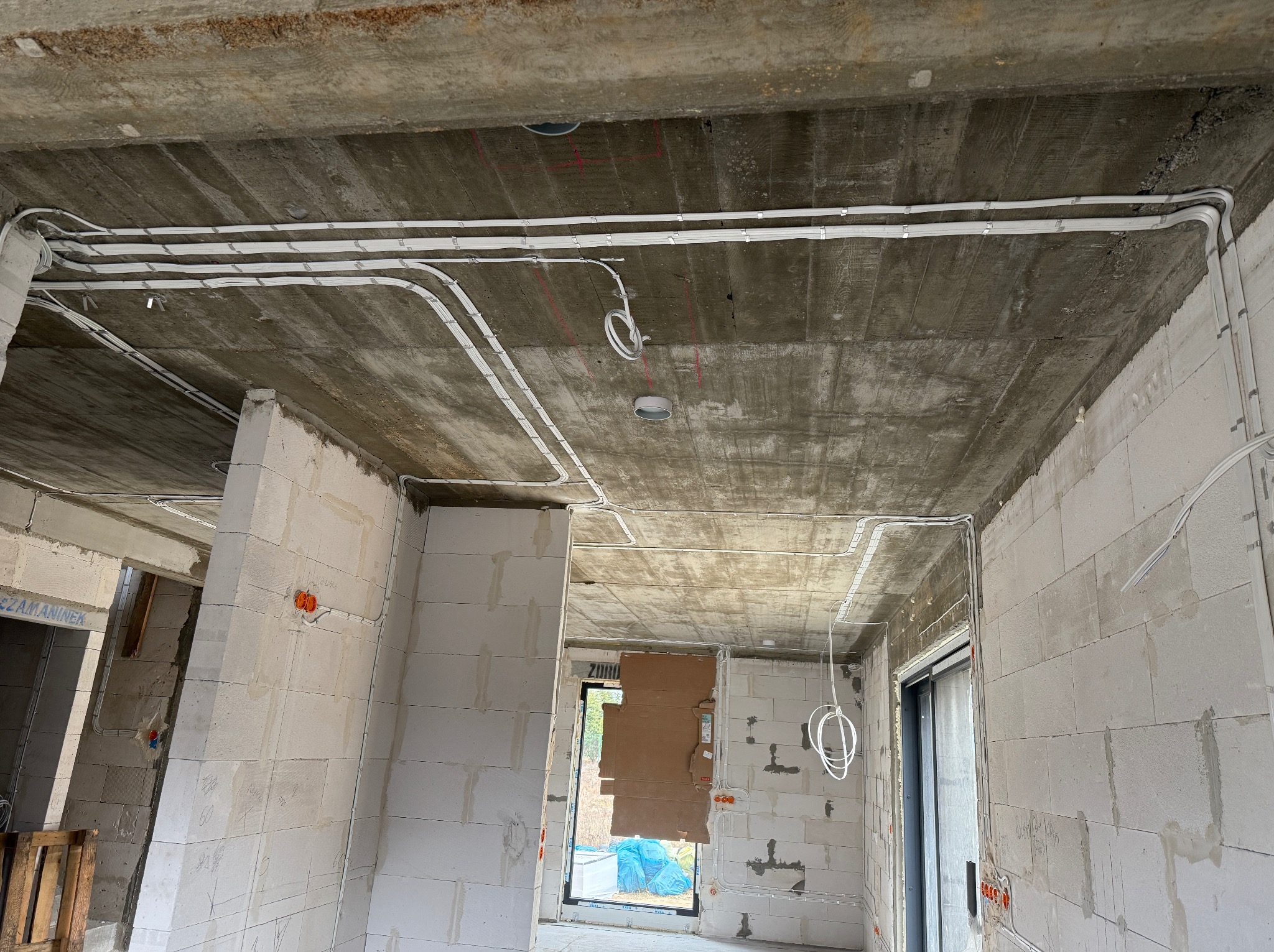 Surowy strop z widocznymi białymi kablami elektrycznymi, zamocowanymi do betonu. W tle ściany z bloczków i otwory okienne. Instalacja w trakcie montażu.