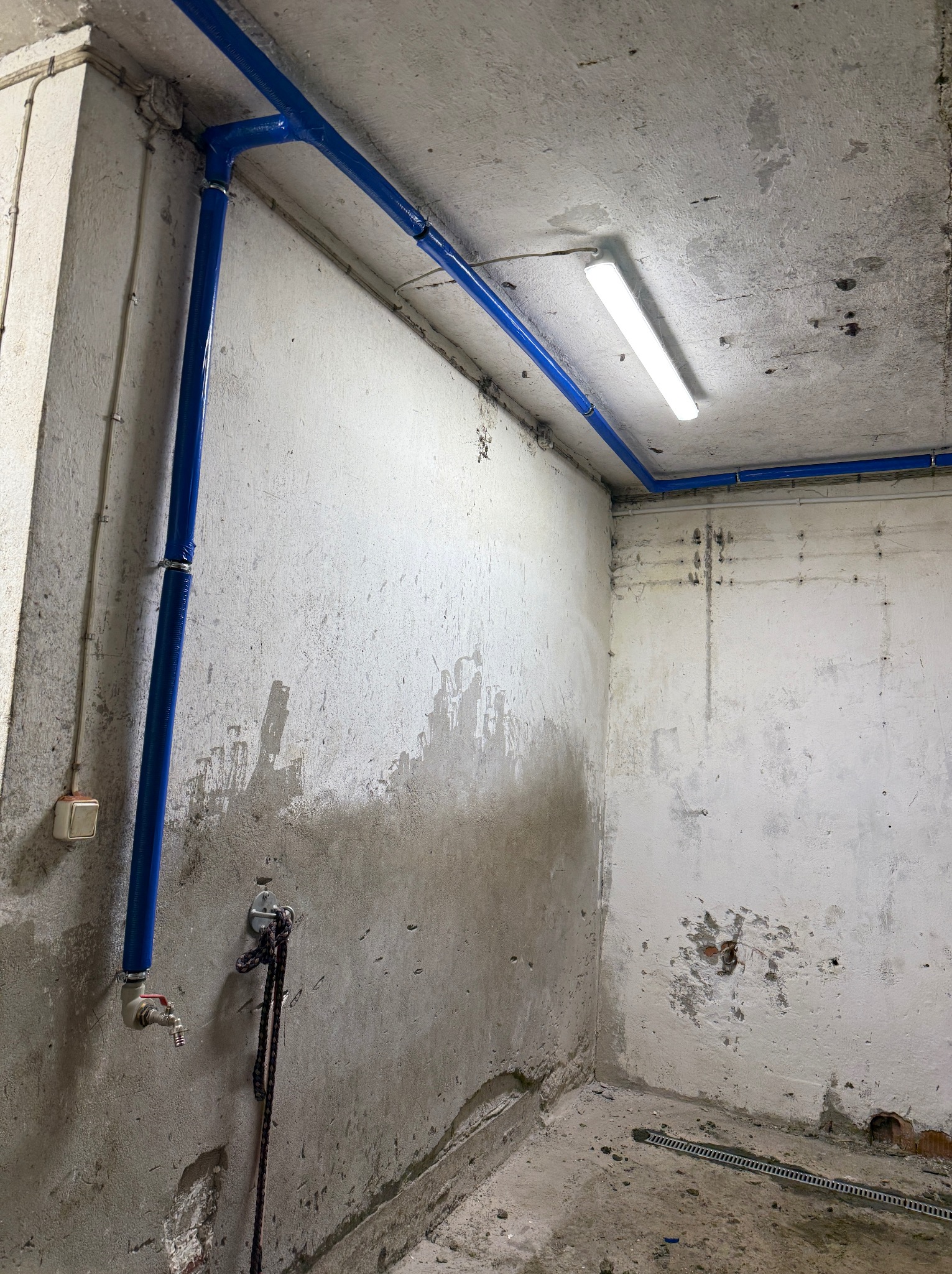 Niebieskie rury instalacji wodnej prowadzone wzdłuż sufitu i ściany w surowym pomieszczeniu z lampą. Widoczny kran i fragment odpływu liniowego w podłodze.