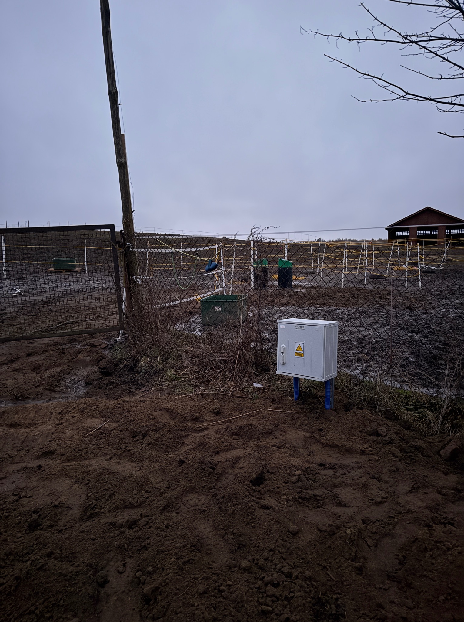 Zewnętrzna skrzynka elektryczna z ostrzeżeniem o napięciu, zamontowana na niebieskich rurach, widoczna na tle błotnistego terenu i ogrodzenia z siatki.