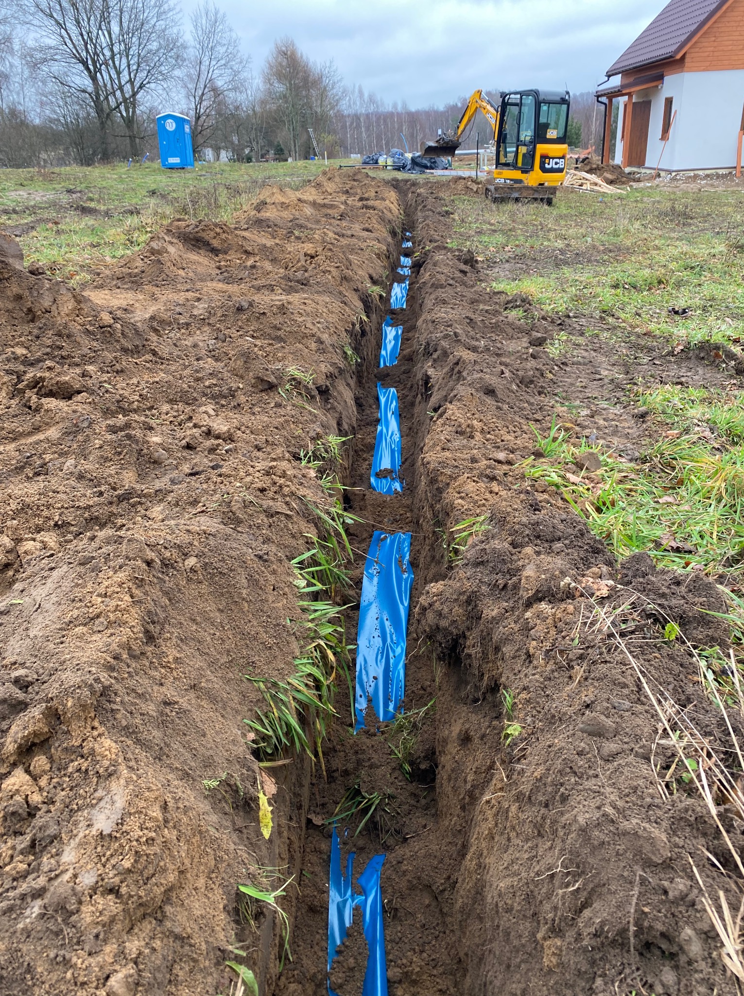 Wykopany rów z niebieskimi rurami w folii ochronnej, na tle koparka JCB, przenośna toaleta i budynek w trakcie budowy.