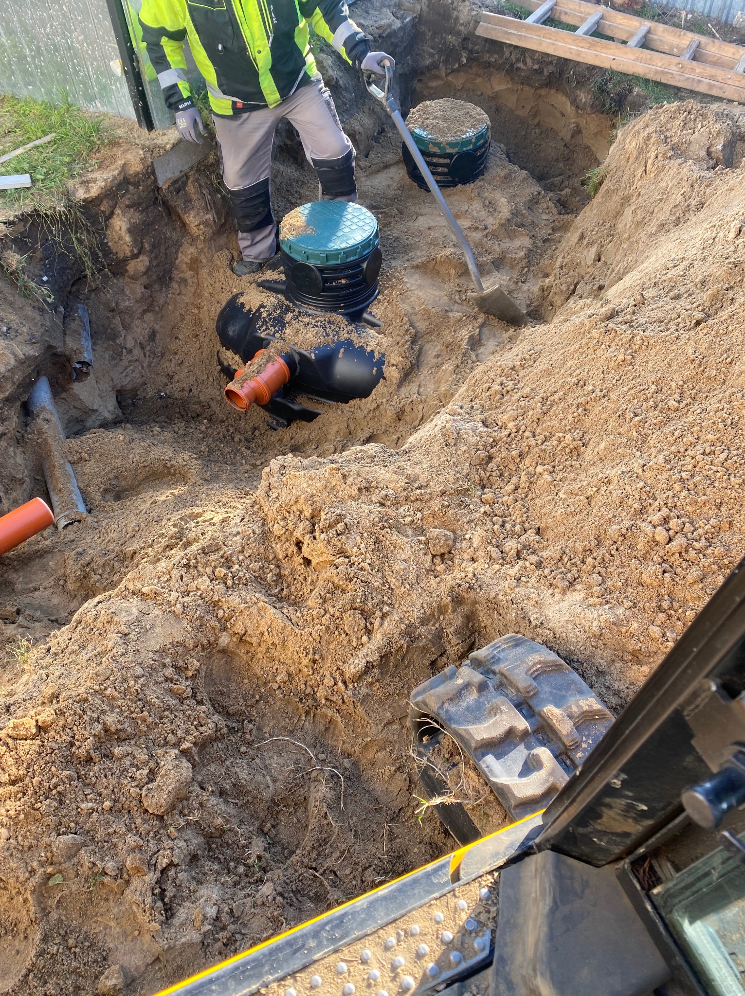 Wykop pod instalacje z widocznymi rurami, studzienkami i fragmentem koparki. Pracownik z łopatą w tle. Ujęcie z góry, ukazujące postęp prac ziemnych.