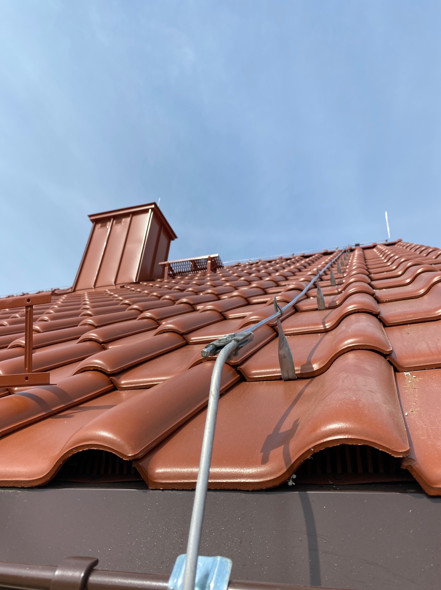 Czerwona dachówka z zainstalowaną instalacją odgromową, widok z dołu na błękitne niebo. Metalowy komin w tle. Specyficzne mocowania instalacji.