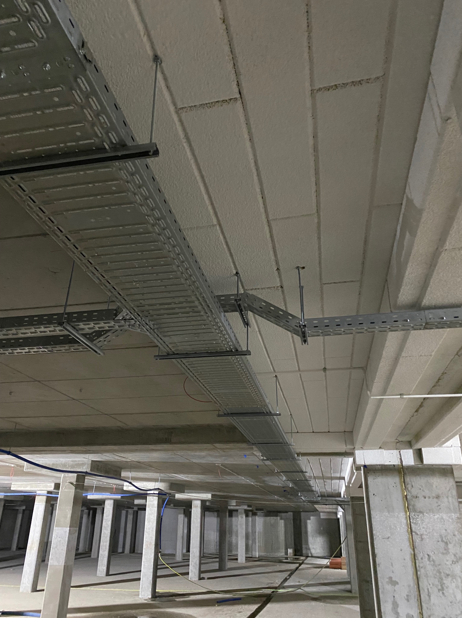Metalowe korytka kablowe pod sufitem, zamocowane na stalowych prętach, widoczne przewody. Surowy stan budynku w trakcie instalacji.