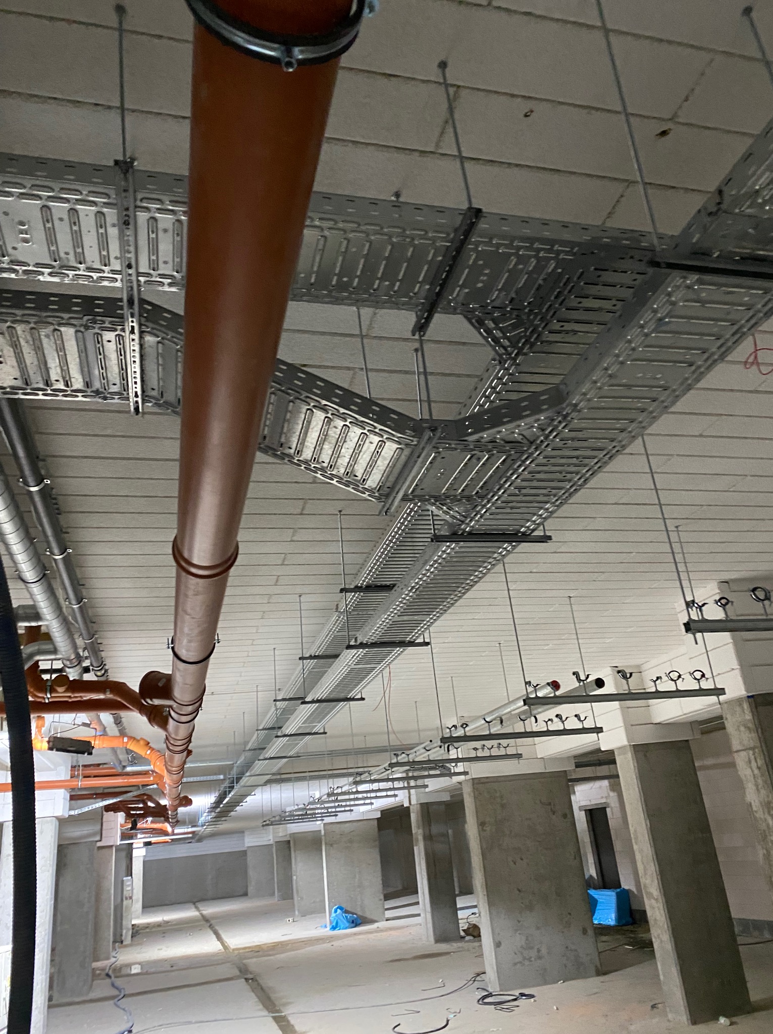 Widok z dołu na sufit z zamontowanymi korytkami kablowymi i rurami wentylacyjnymi w surowym wnętrzu budynku. Szare betonowe słupy w tle.