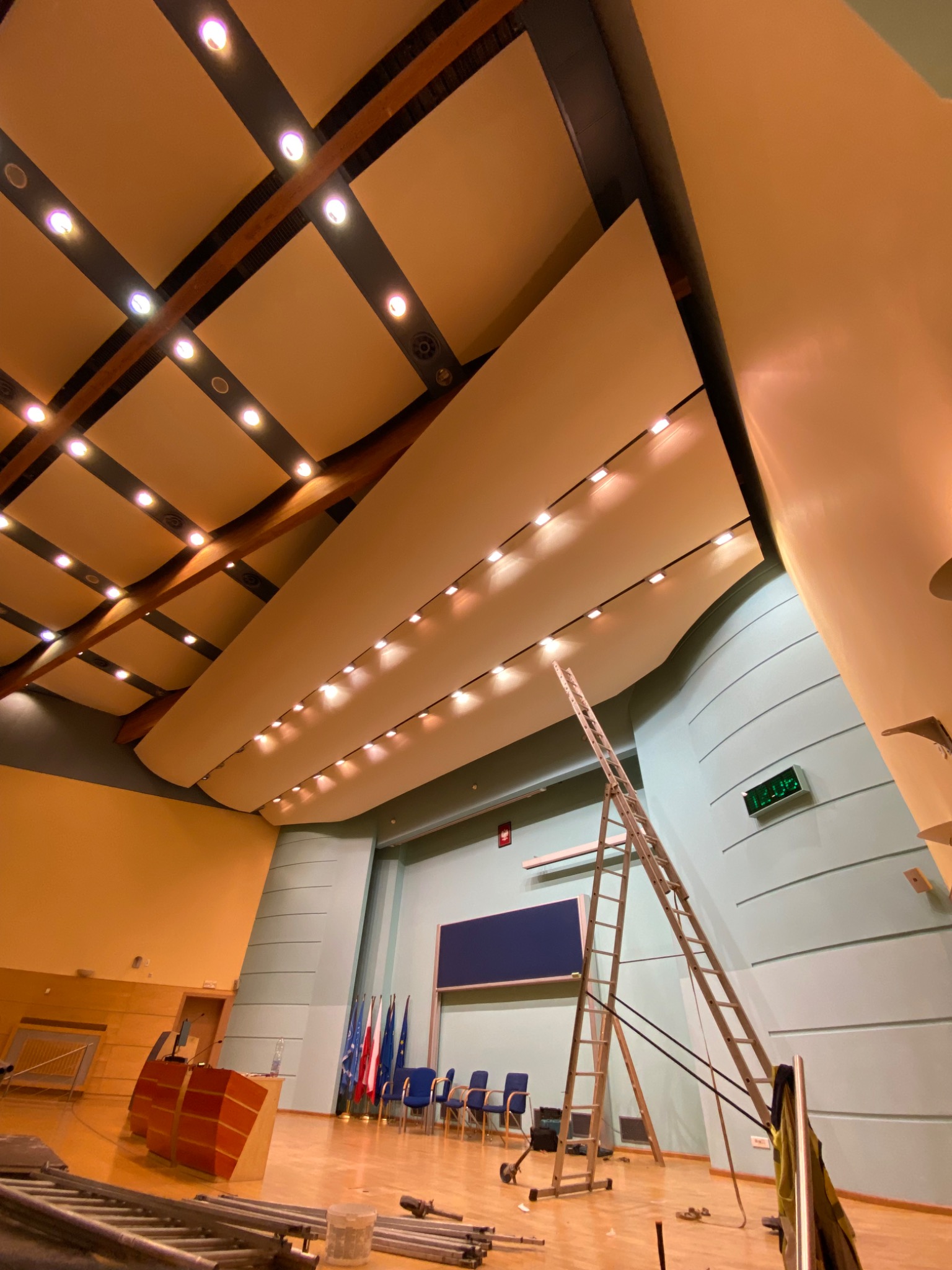 Montaż oświetlenia w sali konferencyjnej z drewnianą konstrukcją sufitu i drabiny. Widoczne flagi państwowe i podium. Oświetlenie punktowe w suficie.