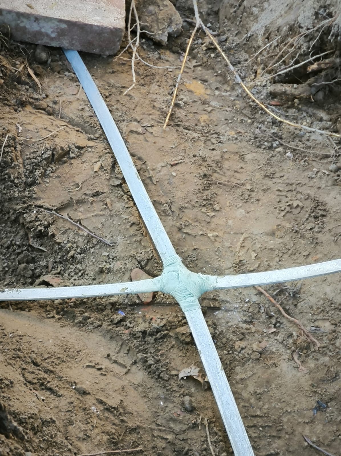 Połączenie przewodów uziemiających w wykopie, zabezpieczone taśmą izolacyjną. Widoczne fragmenty cegieł i korzenie roślin.