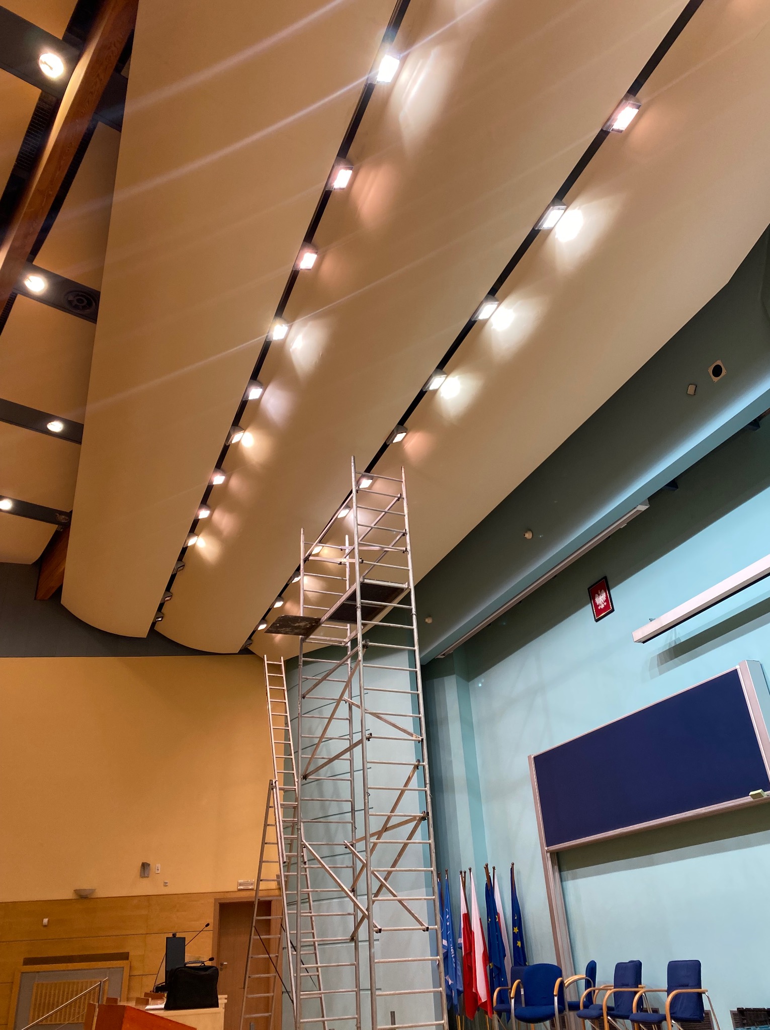 Sala konferencyjna w trakcie montażu oświetlenia sufitowego. Widoczne rusztowanie i drabina, rzędy lamp, flagi i tablica.