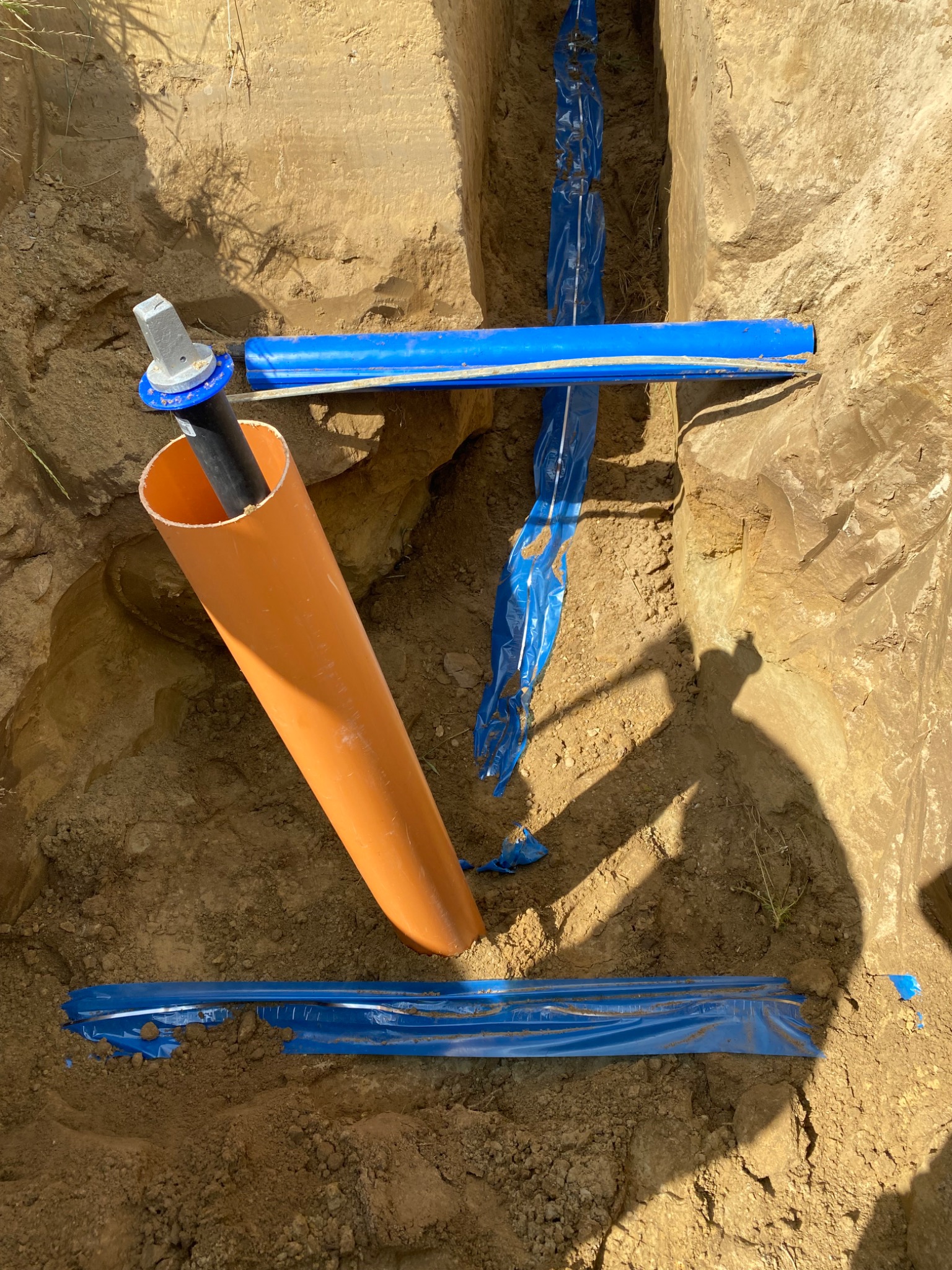 Wykop z rurami: pomarańczowa pionowa, niebieskie poziome z folią ochronną. Widok z góry na instalację w ziemi, przygotowaną do zasypania.