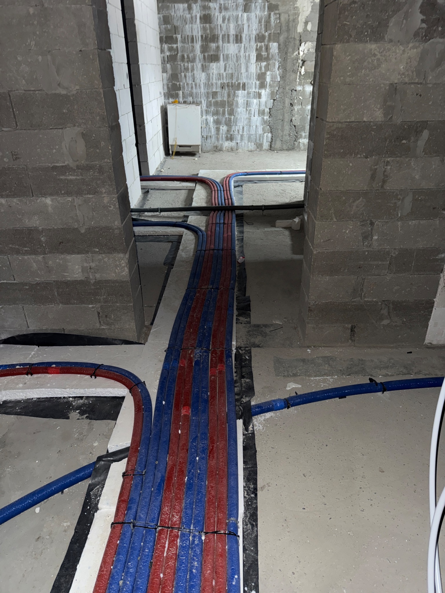 Ułożone rury ogrzewania podłogowego w kolorze niebieskim i czerwonym, prowadzone wzdłuż ściany z pustaków, na podłożu z izolacją. Widok z góry, surowy stan.