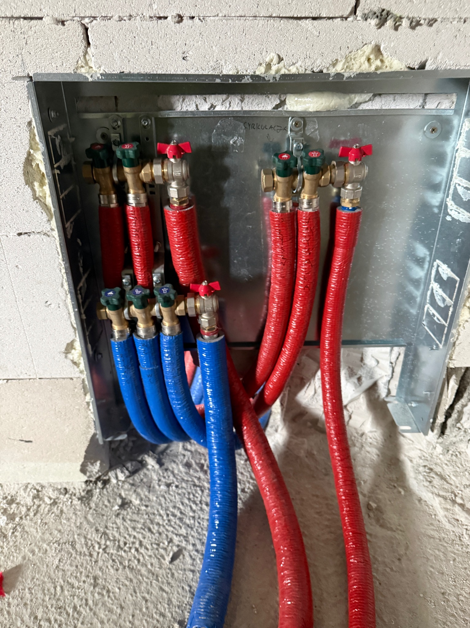 Zabudowa z zaworami i rurami w izolacji: czerwone do ciepłej wody, niebieskie do zimnej. Widoczne ślady montażu na ścianie z betonu komórkowego.