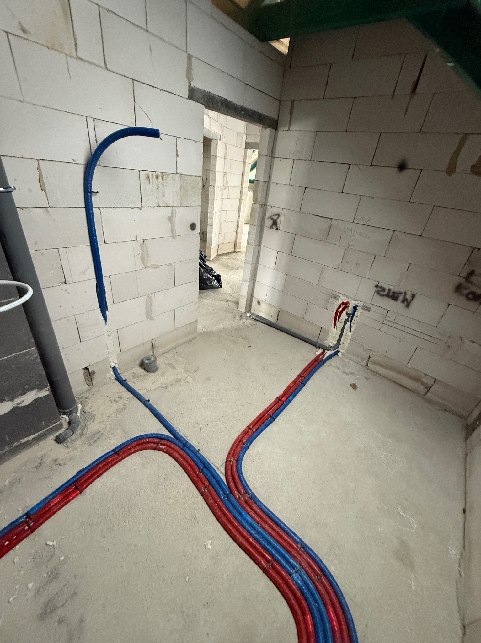 Wnętrze w trakcie budowy z widocznymi, ułożonymi na podłodze rurami instalacji wodnej w kolorach czerwonym i niebieskim, wychodzącymi ze ściany z białego bloczka.