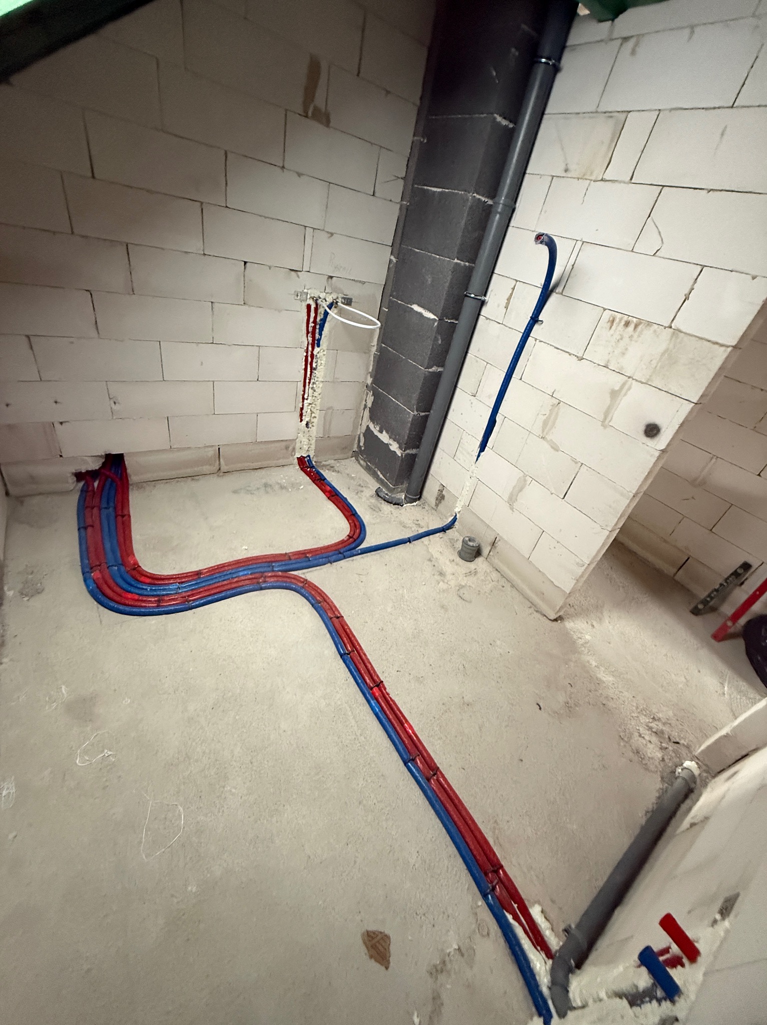 Ułożone rury instalacji wodnej (czerwone i niebieskie) na podłodze w surowym wnętrzu z białych bloczków. Pionowa rura kanalizacyjna w tle. Widoczne pianowanie.