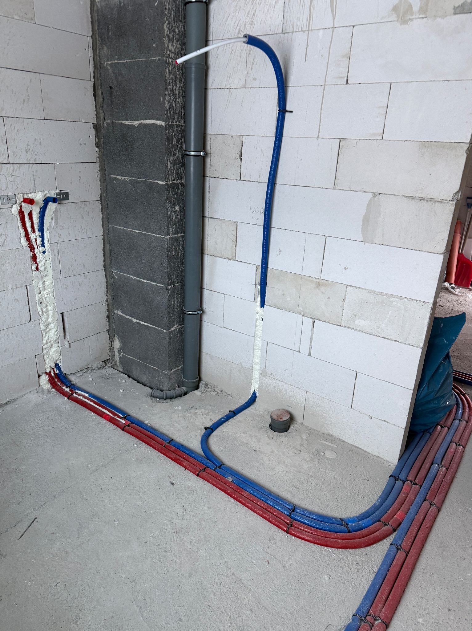 Wnętrze w trakcie prac hydraulicznych: rury grzewcze (czerwone i niebieskie) prowadzone wzdłuż ścian z betonu komórkowego, pion kanalizacyjny i pianka montażowa.