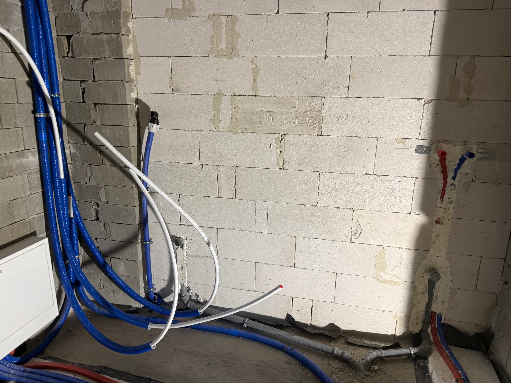 Niebieskie i białe rury instalacji wodnej w surowym wnętrzu z białych bloczków. Część rur ułożona na podłodze, inne przymocowane do ściany. Widoczne napisy na ścianie.