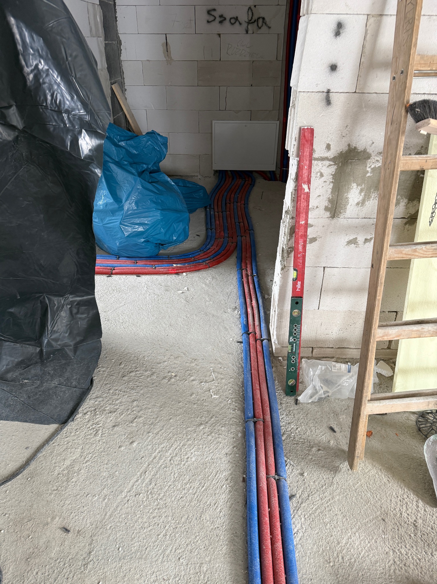 Rury ogrzewania podłogowego w kolorze czerwonym i niebieskim, ułożone na podłodze w surowym stanie, z poziomnicą i drabiną w tle, w trakcie instalacji.