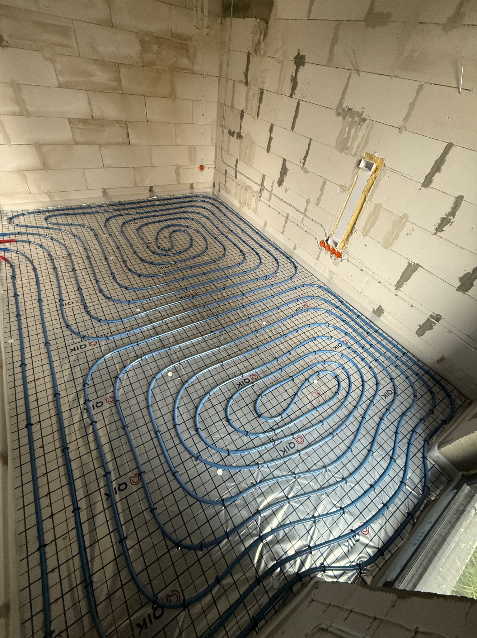 Niebieskie rury ogrzewania podłogowego ułożone na folii i siatce w surowym wnętrzu z białych bloczków. Widoczne pętle rur i instalacja elektryczna w ścianie.