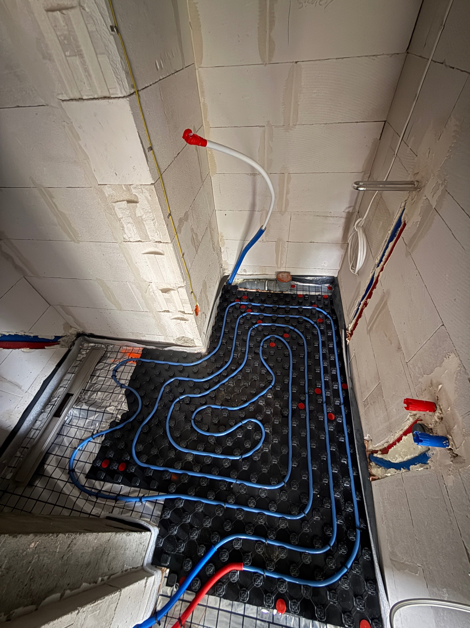 Niebieskie rury ogrzewania podłogowego ułożone na czarnej macie w surowym wnętrzu z białych bloczków, widoczne elementy instalacji wodnej i elektrycznej.