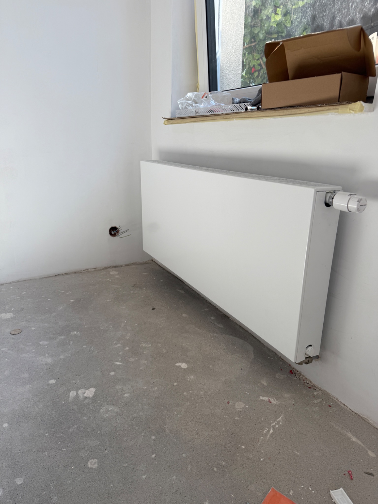 Biały, panelowy grzejnik zamontowany przy ścianie w surowym wnętrzu. Widoczne ślady po pracach wykończeniowych na podłodze i wystające przewody elektryczne.
