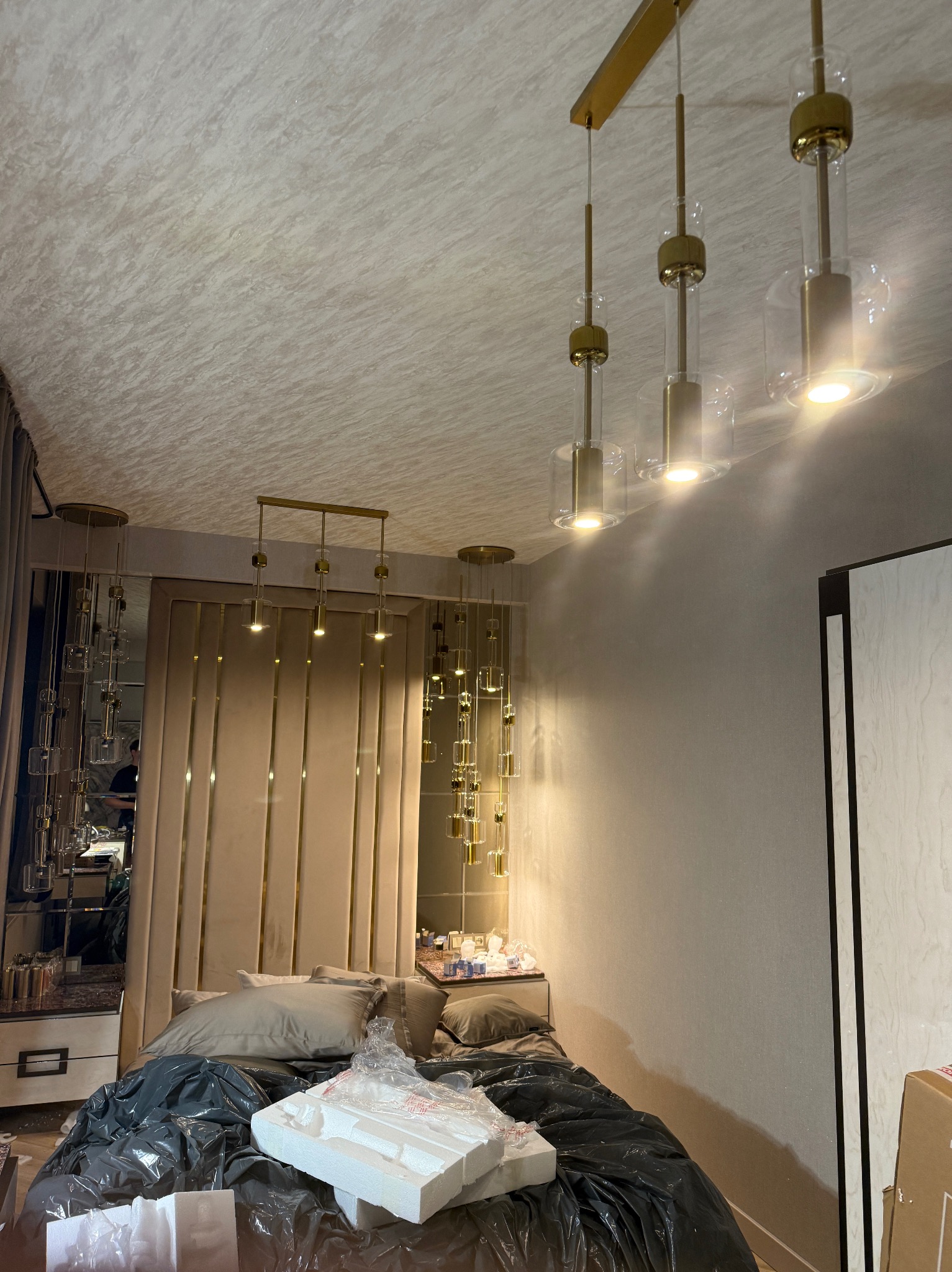 Nowoczesne lampy wiszące z elementami złota nad łóżkiem w sypialni. Widoczne odbicie w lustrze i elementy dekoracyjne. Oświetlenie w stylu glamour.