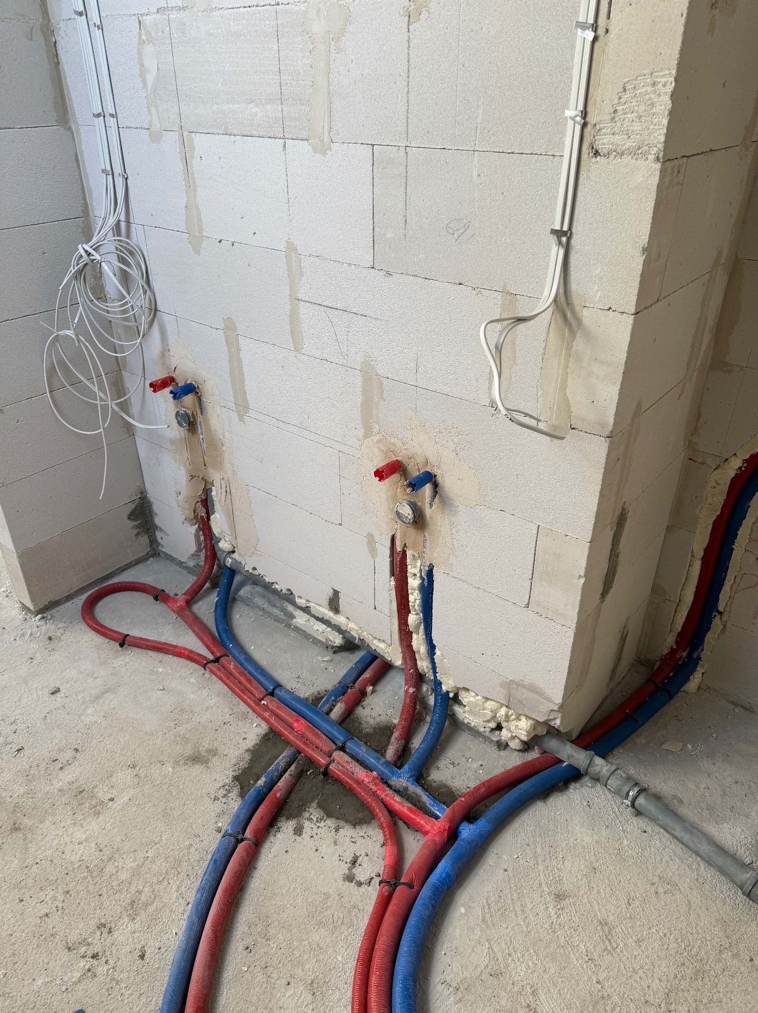 Instalacja wodna i elektryczna w surowym wnętrzu: czerwone i niebieskie rury wodne oraz białe kable elektryczne prowadzone wzdłuż ściany z betonu komórkowego.