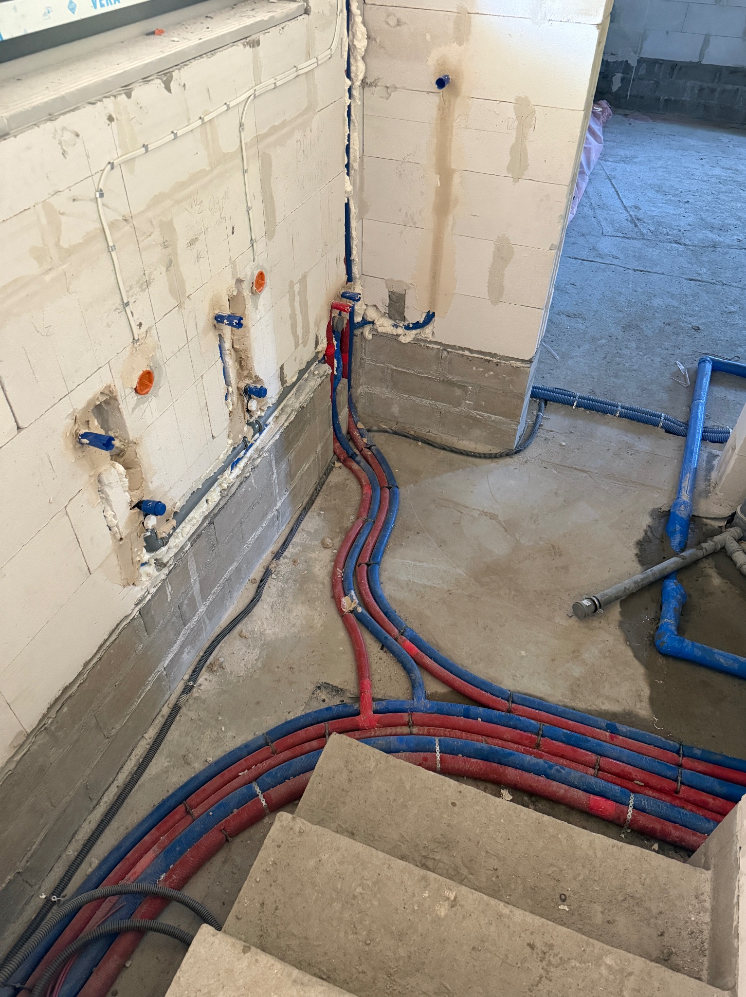Wnętrze w trakcie prac hydraulicznych: czerwono-niebieskie rury prowadzone wzdłuż ściany z betonu komórkowego, z widocznymi punktami przyłączeniowymi.
