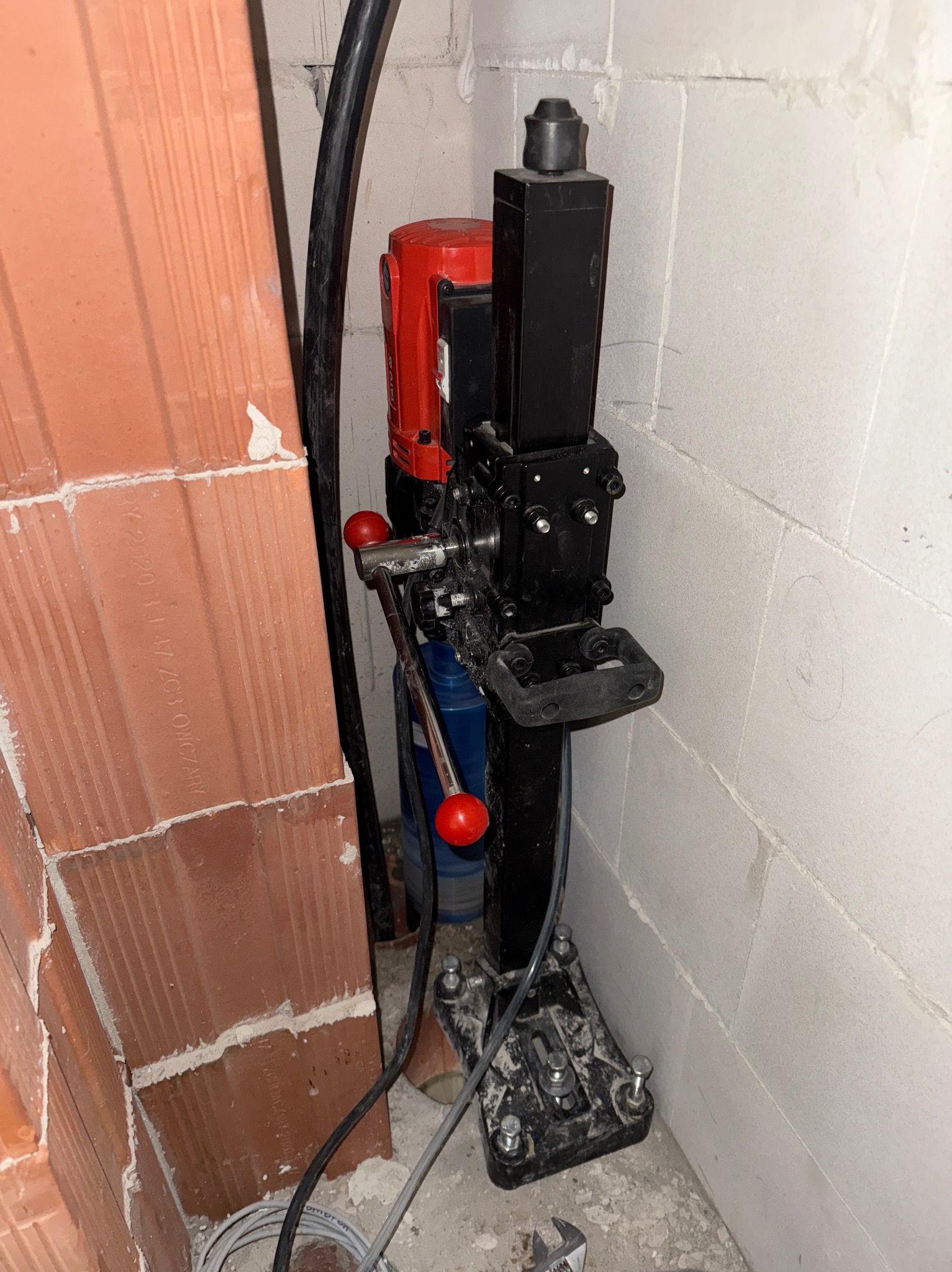 Wiertnica diamentowa z czerwoną obudową silnika, zamocowana do ściany z betonu komórkowego. Widoczny przewód zasilający i fragment ściany z cegły.