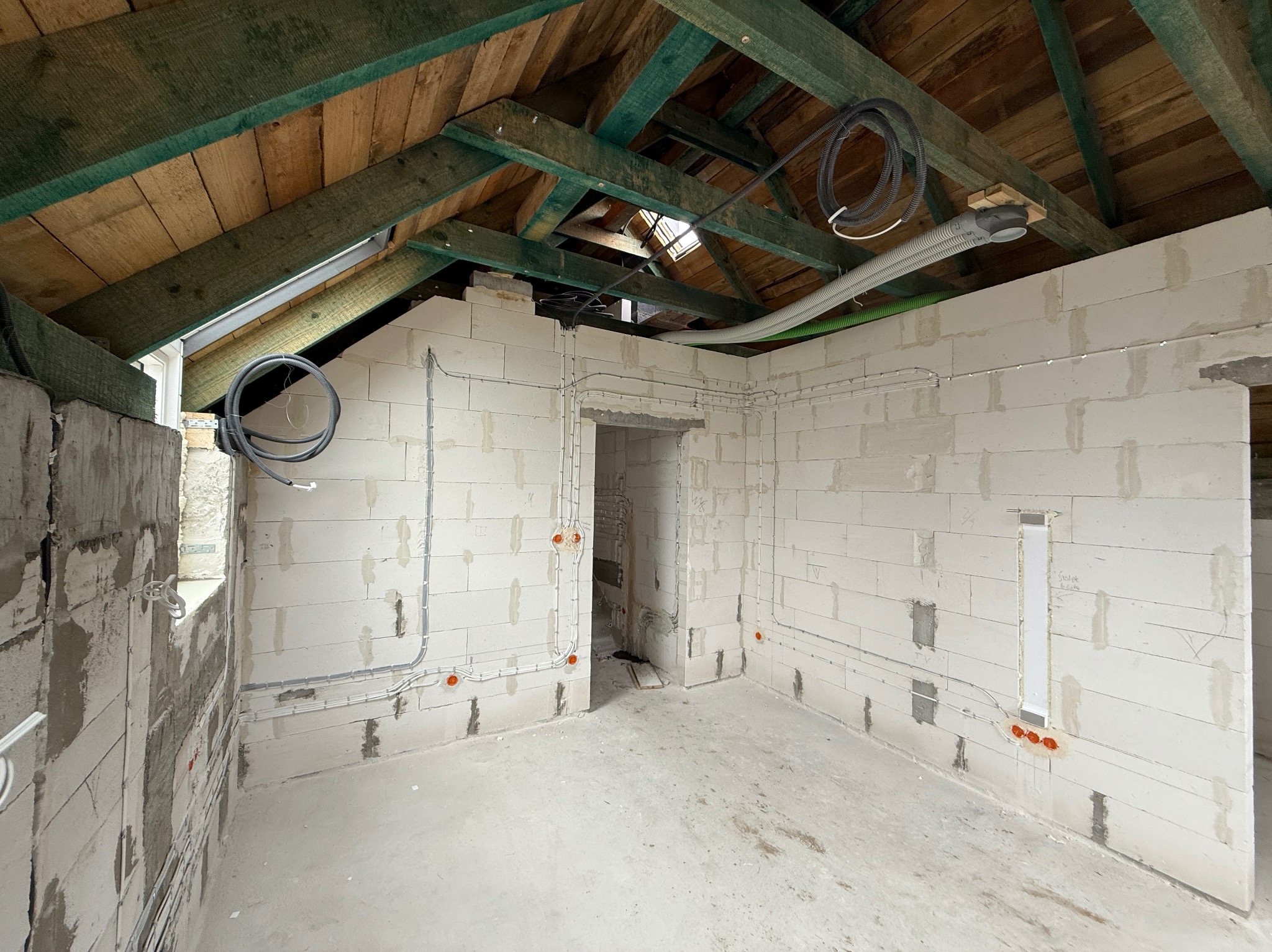 Surowy stan poddasza z widoczną instalacją elektryczną i wentylacyjną na ścianach z betonu komórkowego oraz drewnianą konstrukcją dachu. Widoczne kable i rury.