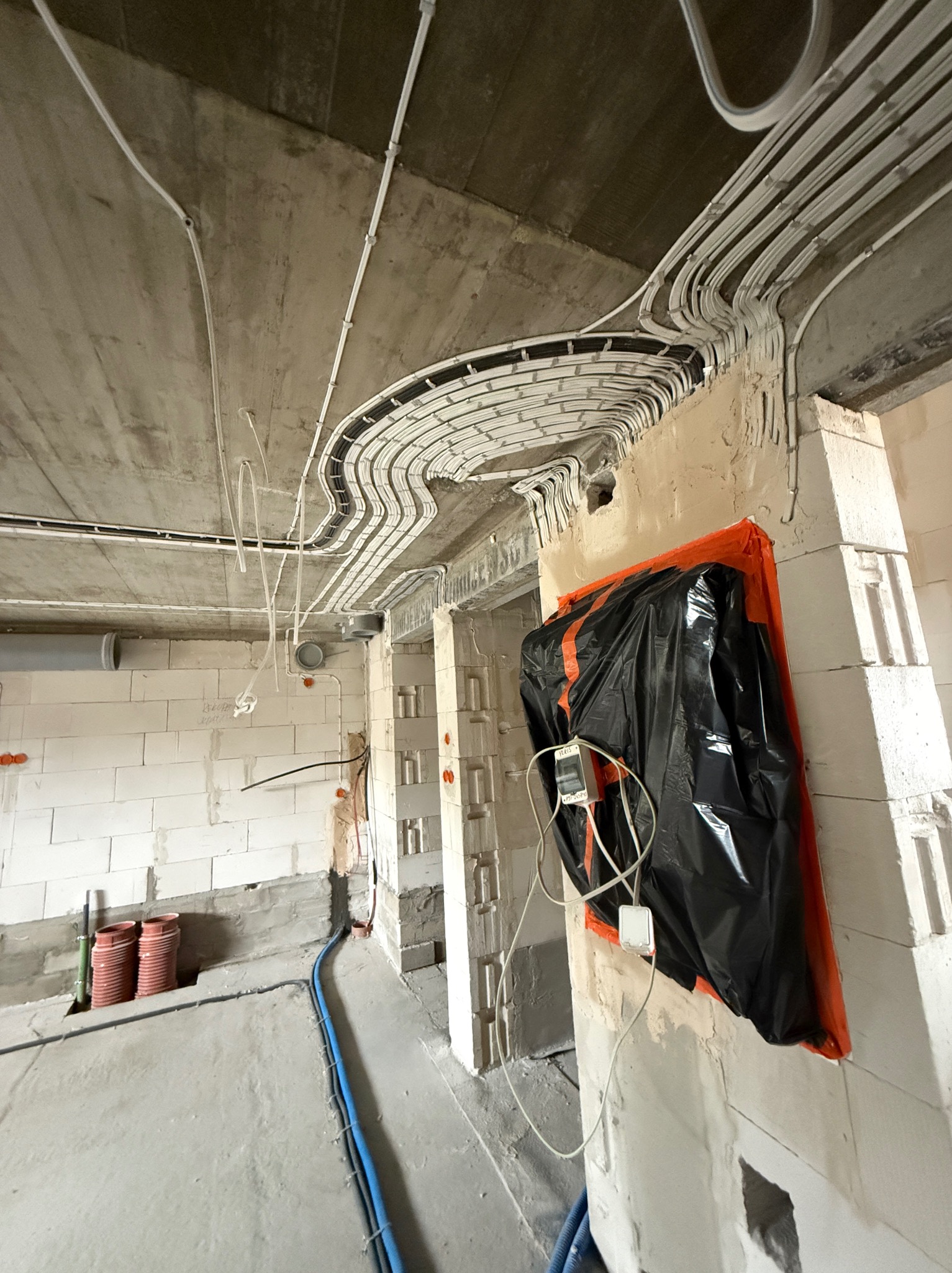 Wnętrze w trakcie prac wykończeniowych: widoczne liczne kable elektryczne ułożone pod sufitem, ściany z białego bloczka, zabezpieczony element instalacji folią.