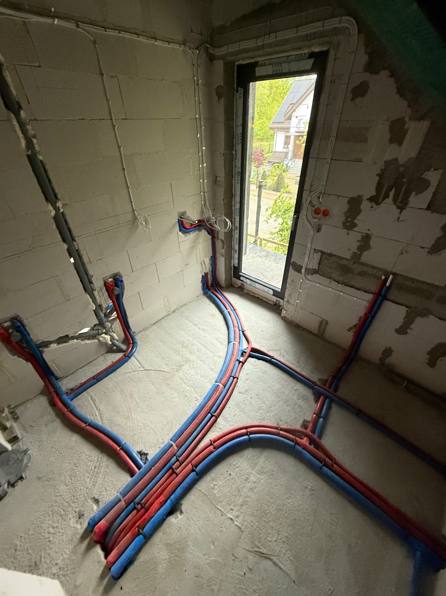 Surowe wnętrze z instalacją hydrauliczną: czerwono-niebieskie rury prowadzone po podłodze i ścianach, widoczne okno z zielenią na zewnątrz, ściany w stanie surowym.