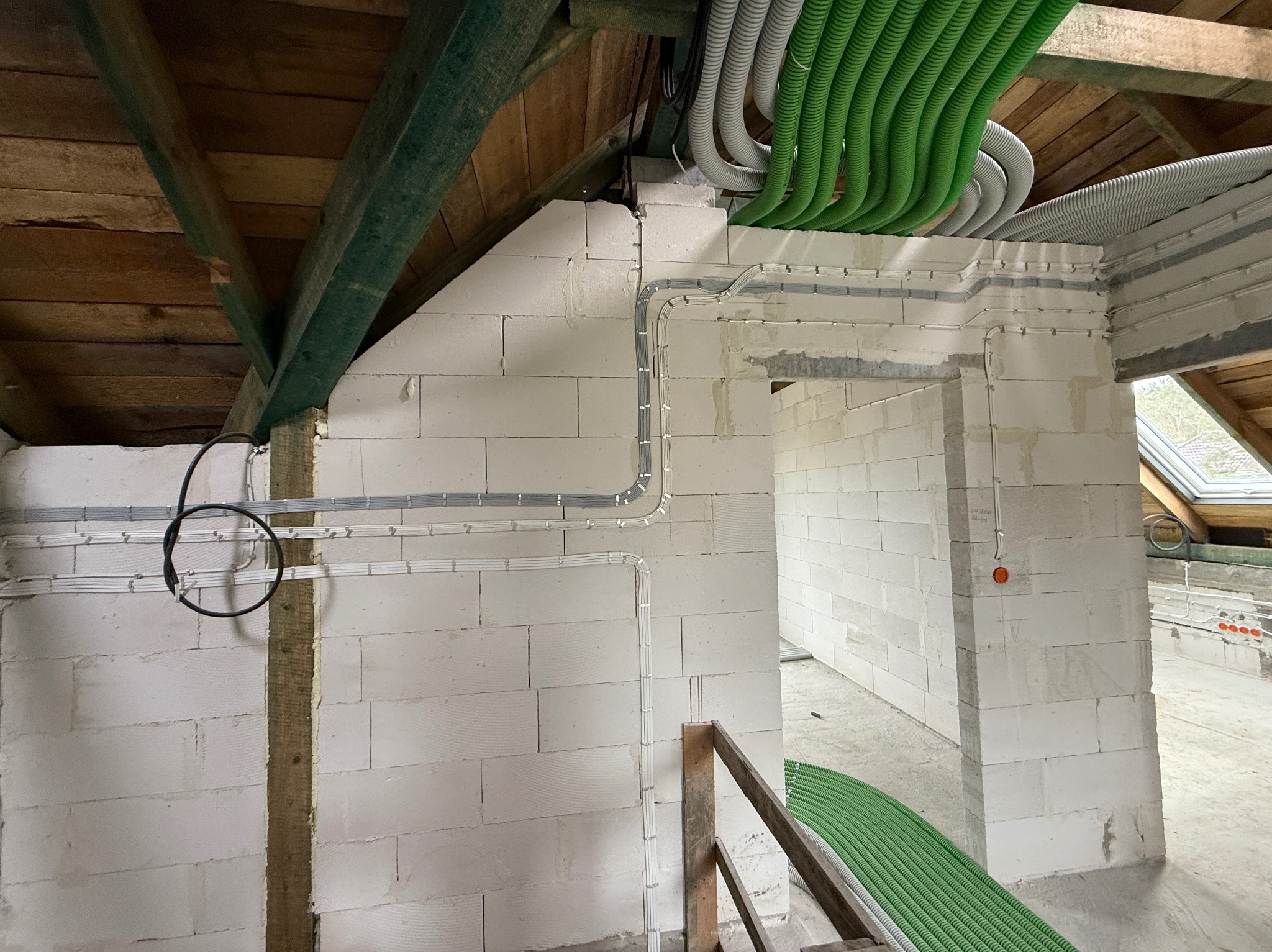 Instalacja elektryczna i hydrauliczna w surowym wnętrzu poddasza. Widoczne kable w peszlach i rury, ściany z białego bloczka, drewniana konstrukcja dachu.