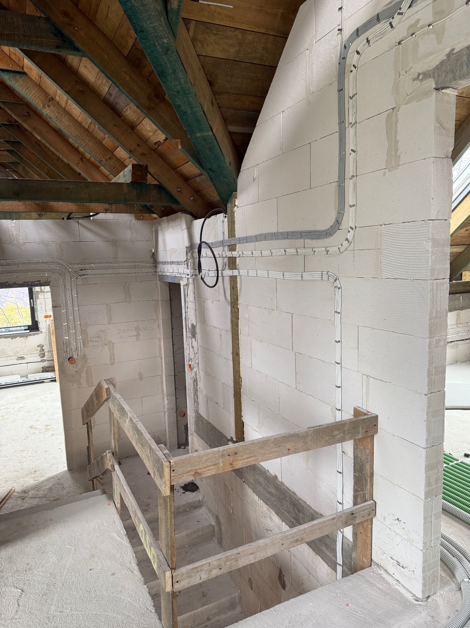 Wnętrze w trakcie prac wykończeniowych: białe ściany z betonu komórkowego, widoczne kable elektryczne prowadzone po ścianach, drewniana konstrukcja dachu i schody.