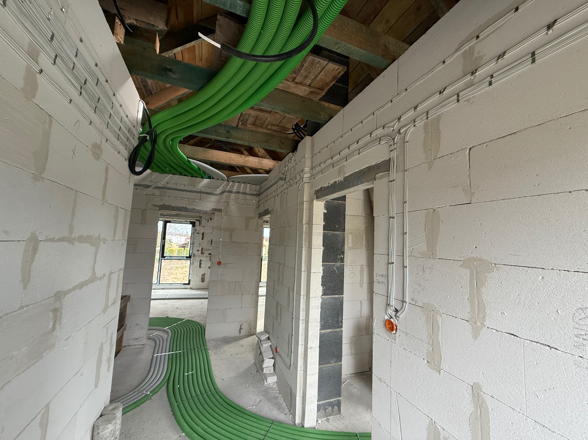Wnętrze w stanie surowym z rozprowadzoną instalacją elektryczną (białe kable) i ogrzewaniem podłogowym (zielone rury) w Chmielnie. Widok na sufit z drewnianą konstrukcją.