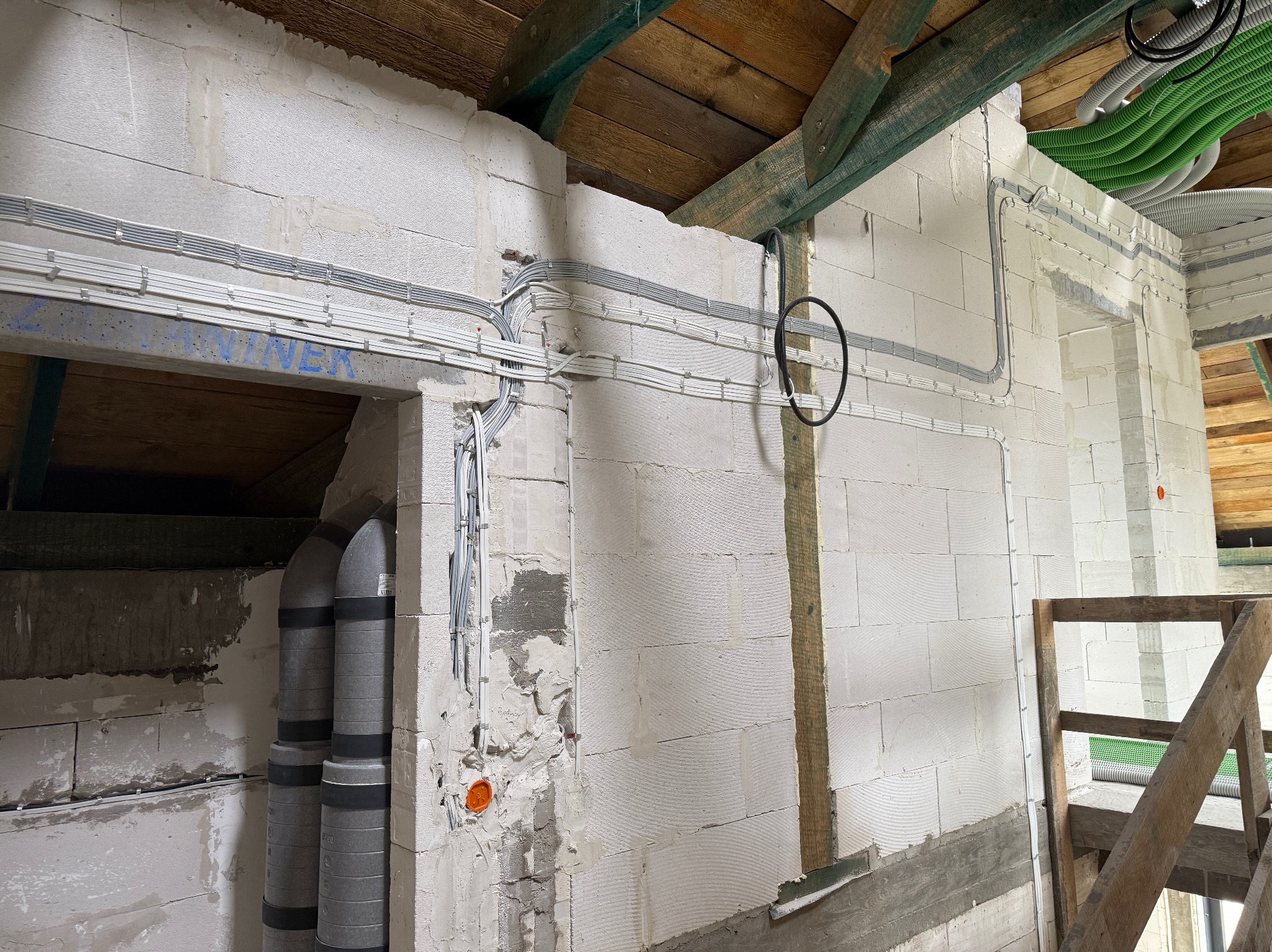 Białe ściany z betonu komórkowego w trakcie budowy, z widocznymi szarymi przewodami elektrycznymi i zielonymi rurami wentylacyjnymi pod drewnianą konstrukcją dachu.